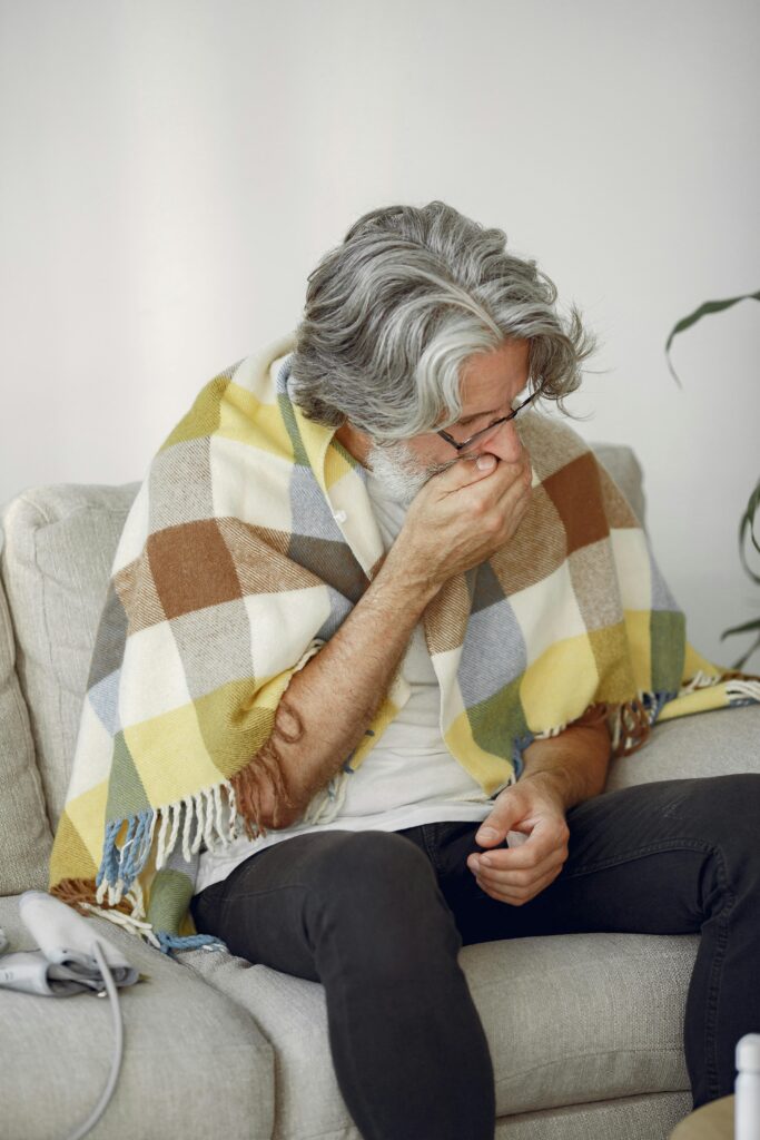 Middle-aged man coughing while sitting on sofa, wrapped in a checkered blanket, at home.